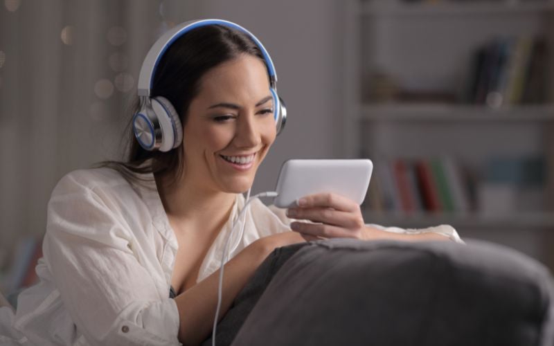 Smiling woman wearing a white shirt and headphones looking at her phone and listening to music while sitting on her couch