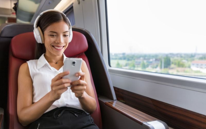 Smiling woman wearing white sleeveless blouse and headphones smiling while listening to music on her phone on a train